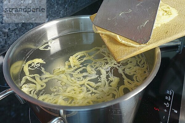 Schwäbische Küche  Zubereitung handgeschabte Spätzle für Spätzle-Auflauf  dreierlei Spätzle  Spätzle klassisch  Mehlspeise  rohen Teig in kochendes Wasser schaben  Spätzleschaber  Spätzlebrett  Kochtopf  Indiktionsherd  Ceranfeld  typisch Schwäbisch  Traditionsküche  vegetarisch  Foodfotografie  Studio  Deutschland  Europa