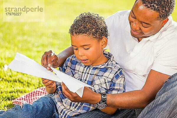 Glücklicher afroamerikanischer Vater und gemischtrassiger Sohn spielen mit Papierflugzeugen im Park