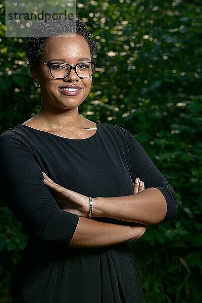 Stehende hübsche selbstbewusste junge Afro Amerikanische Frau mit kurzen Haaren  einer großen Brille in einem schwarzen Kleid vor einem grünen Busch im Sonnenlicht