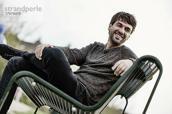 Lachender Junger Mann draußen auf einer Parkbank sitzend  Portrait  Köln  Nordrhein-Westfalen  Deutschland  Europa