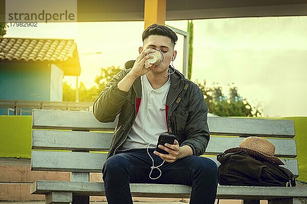 Entspannter attraktiver Junge  der auf einer Bank Musik hört  Junge sitzt auf einer Bank und trinkt Kaffee