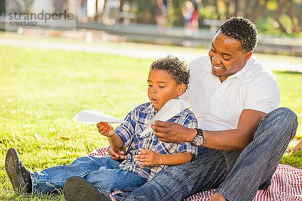 Glücklicher afroamerikanischer Vater und gemischtrassiger Sohn spielen mit Papierflugzeugen im Park