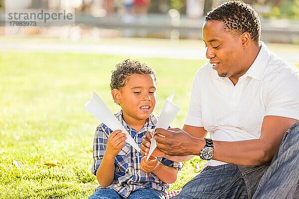 Glücklicher afroamerikanischer Vater und gemischtrassiger Sohn spielen mit Papierflugzeugen im Park