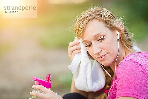 Junge fitte erwachsene Frau im Freien mit Handtuch und Wasserflasche in Trainingskleidung  die mit Kopfhörern Musik hört