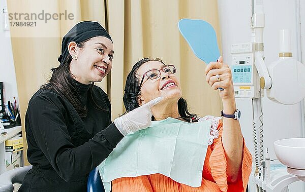 Zufriedene weibliche Klientin in der Zahnklinik  die in den Spiegel schaut  Zahnarzt mit Patientin  die in der Praxis in den Handspiegel lächelt  weibliche Patientin  die die Zähne nach der Zahnbehandlung in der Zahnklinik kontrolliert