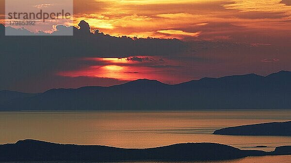 Sonnenuntergang  Sonne groß hinter grauen Wolken und über einem Gebirgszug  Landzungen als Schattenriss im Vordergrund  Meer ruhig  Himmel orangerot  Palaiopolis  Insel Andros  Kykladen  Griechenland  Europa