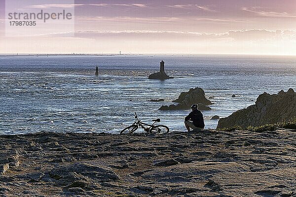 Radfahrer sitzt an Felsenküste  macht Pause  Pointe du Raz  Phare de la Vieille  Silhouette vor Abendhimmel  Cap Sizun  Département Finistère  Bretagne  Frankreich  Europa