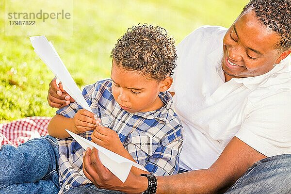 Glücklicher afroamerikanischer Vater und gemischtrassiger Sohn spielen mit Papierflugzeugen im Park