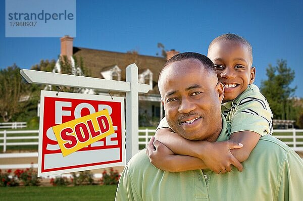 Glücklicher afroamerikanischer Vater und Sohn vor einem neuen Haus und einem verkauften Immobilienschild