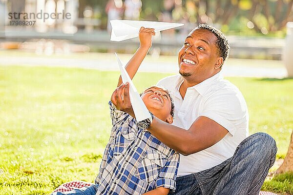 Glücklicher afroamerikanischer Vater und gemischtrassiger Sohn spielen mit Papierflugzeugen im Park