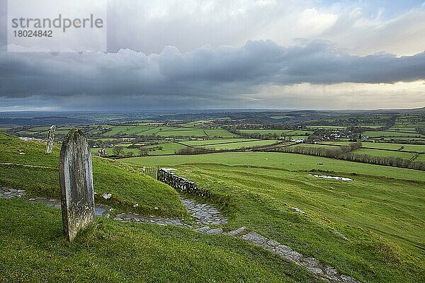 Ansicht des Friedhofs und des Weges  der von der Kirche aus dem 13. Jahrhundert auf Moorland bei Sonnenaufgang führt  mit Regenwolken über der fernen Landschaft  Kirche St. Michael  Brent Tor  Dartmoor N.P.  Devon  England  Dezember