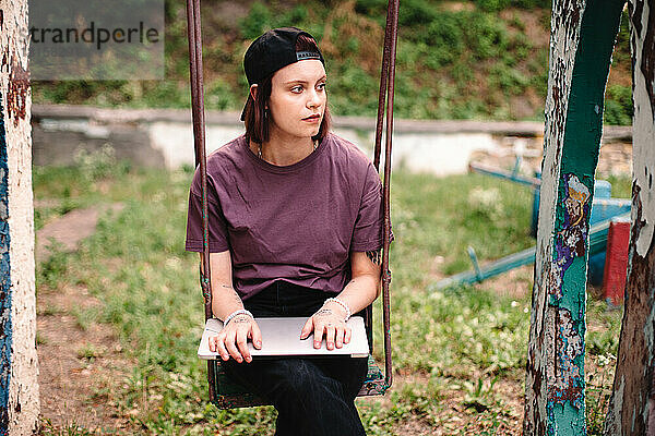 Junge Frau mit Laptop auf einer Schaukel auf einem verlassenen Spielplatz sitzend