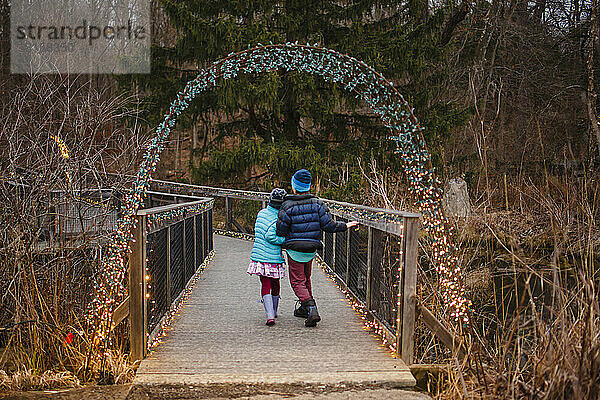 Zwei Kinder gehen auf einen mit Weihnachtsbeleuchtung beleuchteten Weg durch den Wald