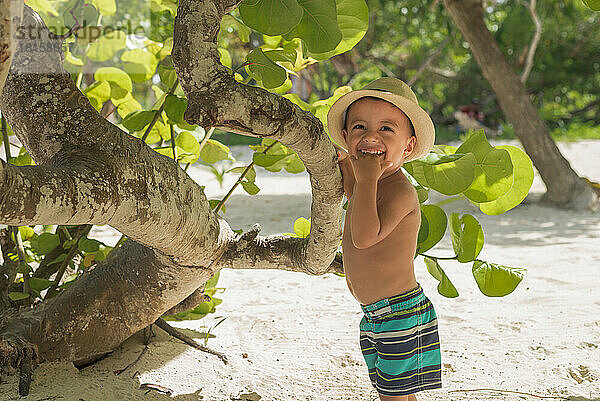 Kleiner Junge mit Hut  der mit Shorts an einem Baum am Strand spielt