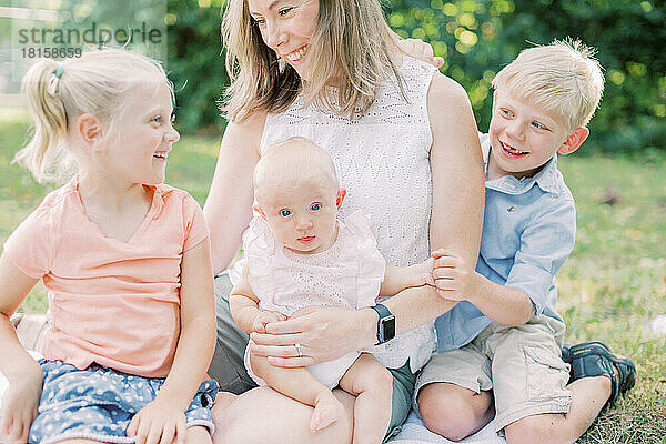 Eine blonde  kaukasische Mutter sitzt mit drei kleinen Kindern zusammen und lächelt sie an.