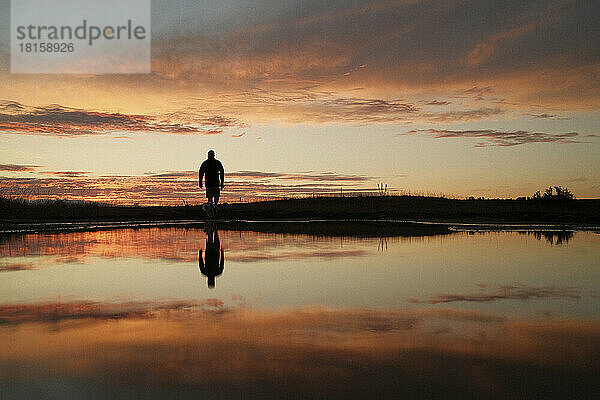 Silhouette einer am Seeufer stehenden Person bei Sonnenuntergang