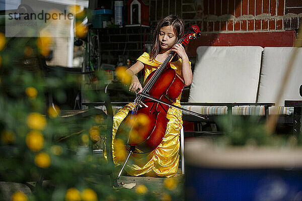 Ein kleines Mädchen im goldenen Kleid spielt Cello im Garten draußen