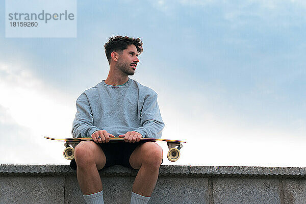Junger Mann sitzt mit einem Skateboard in einem bewölkten Tag bei Sonnenuntergang