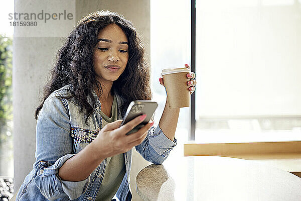 Frau hält Einwegbecher  während sie in einem Café ihr Smartphone benutzt