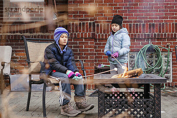Zwei eingemummte Kinder rösten im Winter Marshmallows an einer Feuerstelle