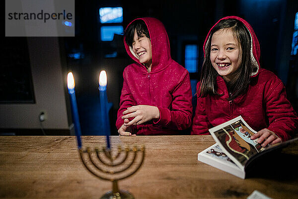 Zwei lachende Kinder sitzen an einem Tisch mit einer beleuchteten Menora zu Hannukah