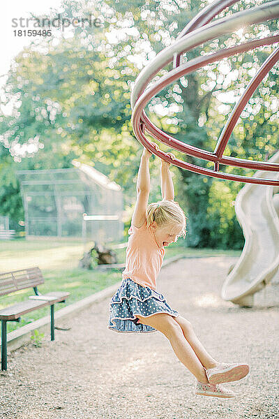 Blondes 4-5-jähriges Mädchen hängt bei Sonnenuntergang an Spielgeräten.