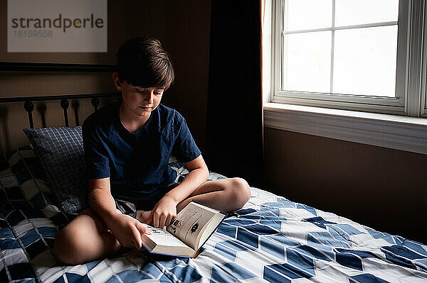 Kleiner Junge liest ruhig ein Buch auf seinem Bett in seinem Schlafzimmer.