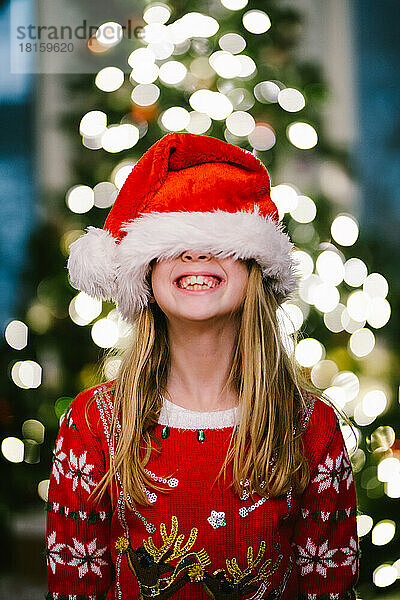 Blondes Kind lächelt mit Weihnachtsmannmütze vor dem Weihnachtsbaum