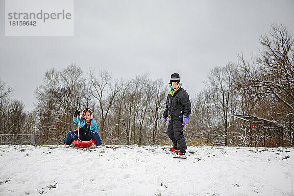 Zwei Kinder spielen auf einem verschneiten Hügel mit Schlitten und Snowboard