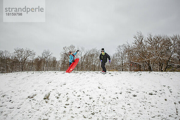 Zwei Kinder stehen auf einem verschneiten Hügel mit einem Schlitten und einem Snowboard