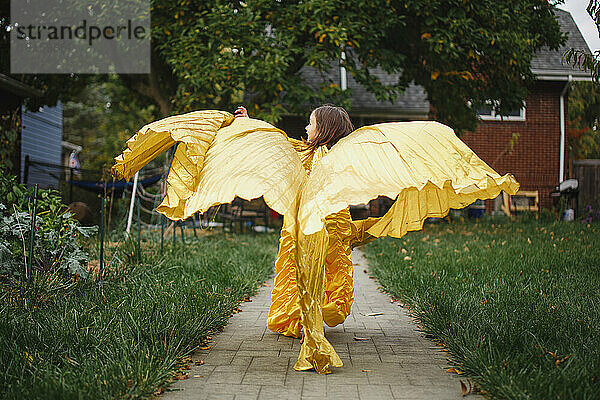Ein kleines Mädchen in goldenem Kleid und mit Flügeln läuft fröhlich durch den Garten