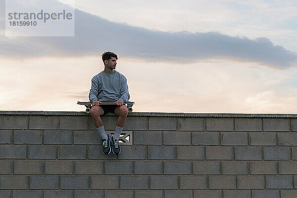 Junger Mann sitzt bei Sonnenuntergang mit einem Skateboard auf einer Mauer
