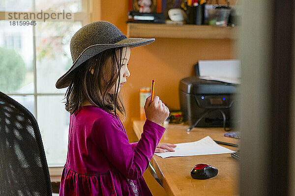 Ein kleines Kind mit Hut sitzt am Schreibtisch mit Papier und Bleistift
