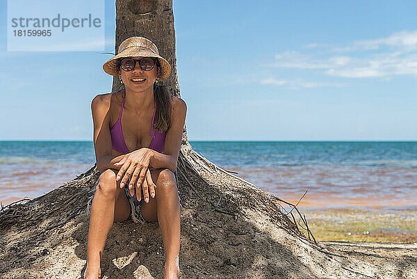Lateinamerikanische Frau unter einer Palme am Strand sitzend und eine Hütte tragend