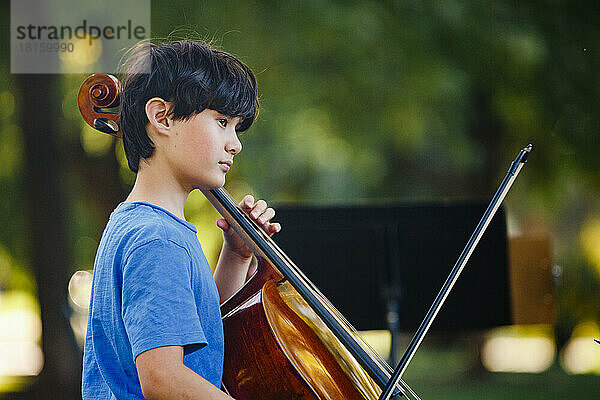 Ein ernster Junge sitzt im sonnenbeschienenen Park und spielt Cello