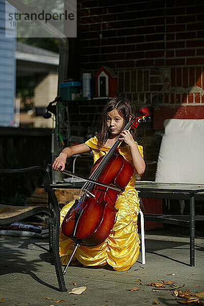 Ein konzentriertes Kind in einem langen goldenen Kleid spielt Cello auf einer Terrasse im Hinterhof