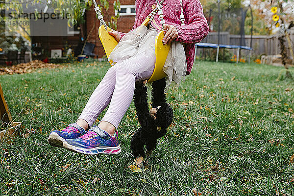 Untere Hälfte eines kleinen Mädchens  das mit einem ausgetopften Affen im Garten schaukelt