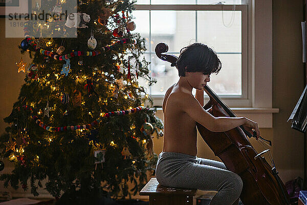 Ein Junge ohne Hemd spielt Cello vor einem beleuchteten Weihnachtsbaum zu Hause