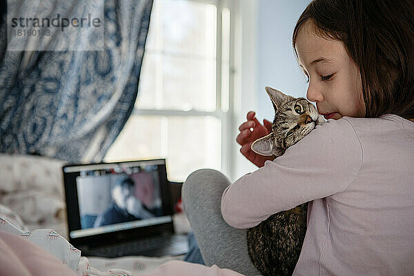 Ein kleines Mädchen sitzt auf dem Bett vor dem Computer und kuschelt mit einem Kätzchen
