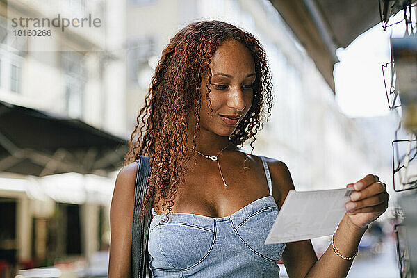 Lächelnde junge Frau mit lockigem Haar  die auf eine Postkarte schaut