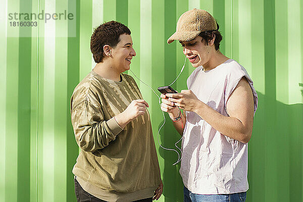 Glückliches nicht-binäres Paar  das sein Smartphone nutzt und mit In-Ear-Kopfhörern Musik hört