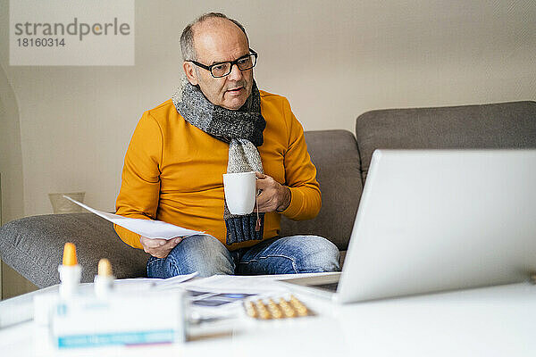 Älterer Mann mit Becher und ärztlichem Bericht blickt im Wohnzimmer auf Laptop