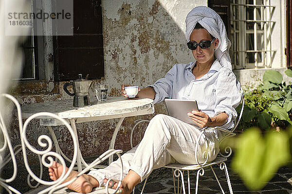 Frau mit Sonnenbrille schaut auf Tablet-PC und trinkt Kaffee auf dem Dach