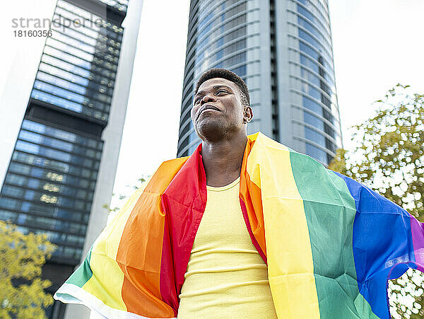 Glücklicher junger Mann in Regenbogenfahne gehüllt vor Wolkenkratzern