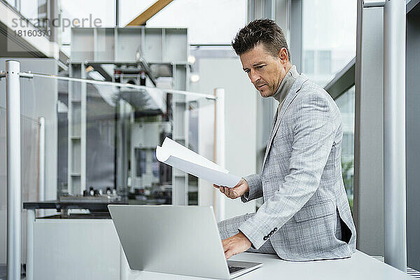 Reifer Geschäftsmann hält Papier mit Laptop am Schreibtisch in der Industrie