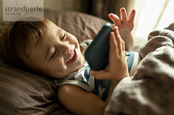 Smiling toddler using mobile phone lying with blanket in bed at home