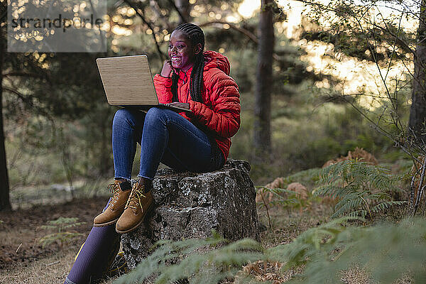 Glückliche junge Frau sitzt mit Laptop auf einem Felsen im Wald