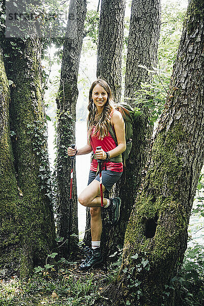 Lächelnde Frau mit Wanderstöcken lehnt an Baumstamm