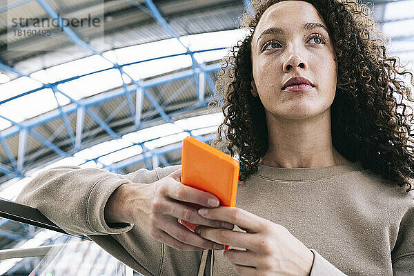 Nachdenkliche Frau mit lockigem Haar hält Mobiltelefon am Geländer