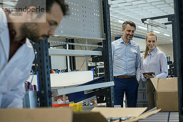 Lächelnder Manager hält Tablet-PC in der Hand eines Geschäftsmannes in der Nähe eines Ingenieurs  der in der Fabrik arbeitet